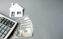 White,Paper,House,With,Dollars,Banknotes,And,Calculator,On,A